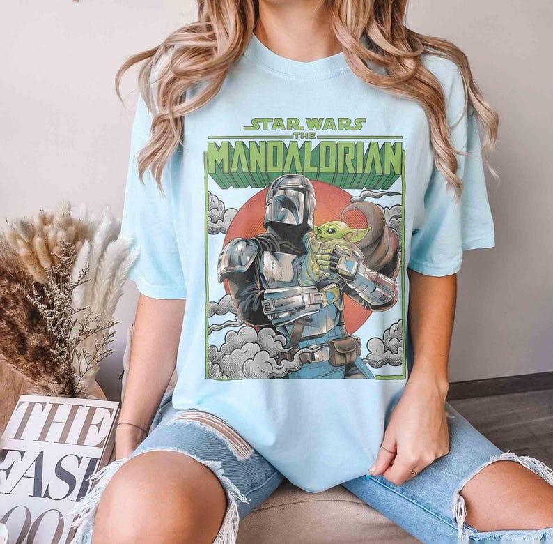 May include: Pale blue t-shirt with a graphic of The Mandalorian and Grogu, with the text "STAR WARS THE MANDALORIAN" in green and white. The design includes a red sun and grey clouds.