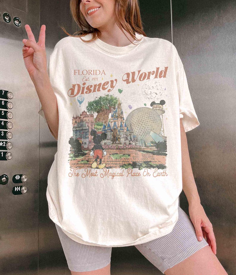 May include: Cream-colored t-shirt featuring a colorful graphic of Disney World landmarks, including Cinderella's castle and Spaceship Earth. The text "Florida Disney World" is printed above the graphic, with the phrase "The Most Magical Place On Earth" below.