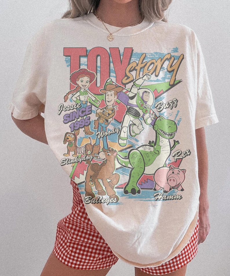 May include: An off-white t-shirt featuring a colorful Toy Story graphic print. The design includes characters Jessie, Woody, Buzz, Slinky Dog, Bullseye, Rex, and Hamm with their names and the text "SINCE 1995" in a distressed, vintage-inspired style.