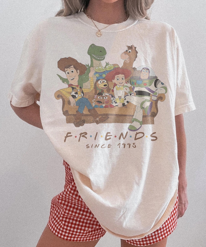May include: An off-white short-sleeve t-shirt featuring a graphic of Toy Story characters sitting on a couch with the text "F.R.I.E.N.D.S SINCE 1995" printed below. The shirt is paired with red and white gingham patterned shorts.