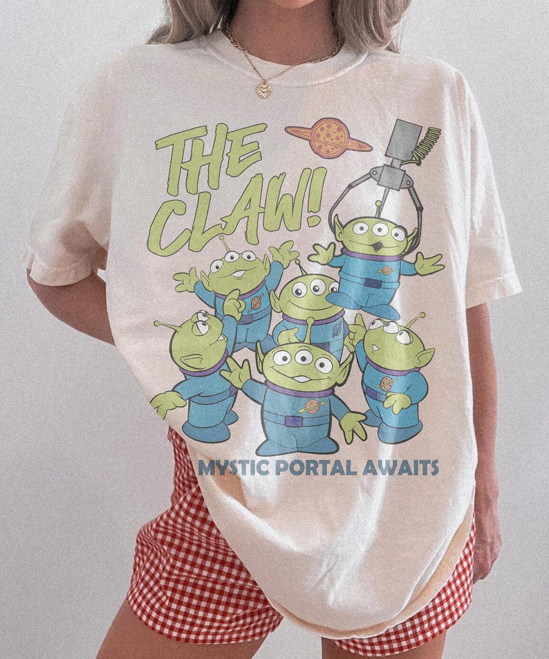 Puede incluir: Una camiseta de manga corta color hueso con un gr&aacute;fico de cinco alien&iacute;genas verdes de tres ojos con trajes espaciales azules. El texto dice THE CLAW! y MYSTIC PORTAL AWAITS. Una garra mec&aacute;nica cuelga sobre los alien&iacute;genas y un planeta.