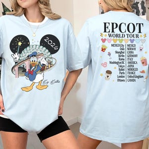 May include: Light blue t-shirt featuring Donald Duck in front of a stylized EPCOT Center design, with the year 2026. The back of the shirt displays "EPCOT WORLD TOUR" and a list of cities. A fun, colorful design.