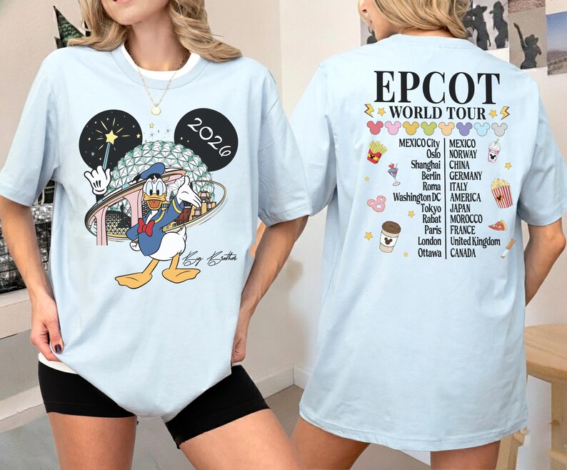 May include: Light blue t-shirt featuring Donald Duck in front of a stylized EPCOT Center design, with the year 2026. The back of the shirt displays "EPCOT WORLD TOUR" and a list of cities. A fun, colorful design.