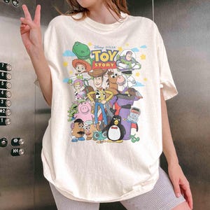 May include: Cream-colored t-shirt featuring a colorful graphic of characters from the Disney Pixar movie "Toy Story." The design includes Woody, Buzz Lightyear, and other beloved characters. The shirt has short sleeves and a relaxed fit.