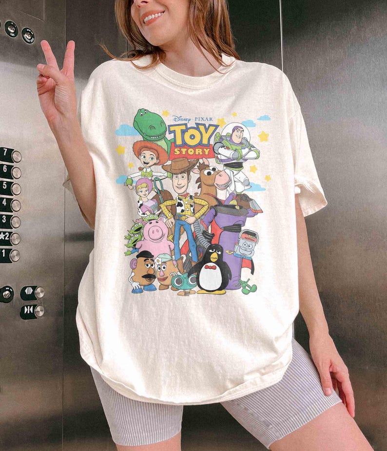 May include: Cream-colored t-shirt featuring a colorful graphic of characters from the Disney Pixar movie "Toy Story." The design includes Woody, Buzz Lightyear, and other beloved characters. The shirt has short sleeves and a relaxed fit.