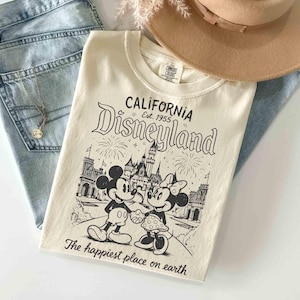 May include: Cream-coloured t-shirt with a black and white graphic of Mickey and Minnie Mouse in front of a castle, with the text "CALIFORNIA Disneyland Est. 1955" and "The happiest place on earth". The shirt is paired with light blue jeans and a tan hat.