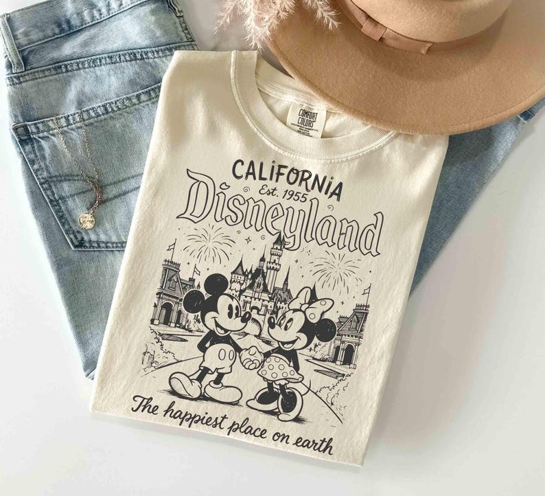 May include: Cream-coloured t-shirt with a black and white graphic of Mickey and Minnie Mouse in front of a castle, with the text "CALIFORNIA Disneyland Est. 1955" and "The happiest place on earth". The shirt is paired with light blue jeans and a tan hat.