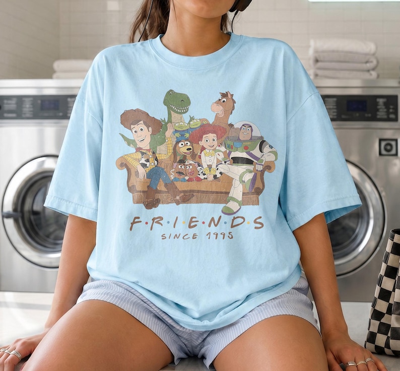 May include: A light blue t-shirt featuring a graphic print of various animated characters sitting on a brown couch. Below the illustration, the text reads "F R I E N D S" in a brown font, with the text "SINCE 1995" printed directly underneath it.