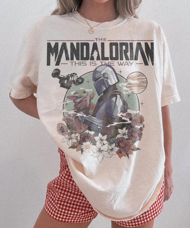 May include: Cream-colored t-shirt featuring a graphic of The Mandalorian and Grogu surrounded by flowers. The text "THE MANDALORIAN - THIS IS THE WAY" is printed across the top. The shirt has short sleeves.