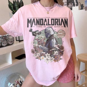 May include: A light pink t-shirt featuring a graphic from The Mandalorian. The design includes the text "THE MANDALORIAN THIS IS THE WAY" above an illustration of the Mandalorian, Grogu, and floral elements. The shirt has short sleeves.