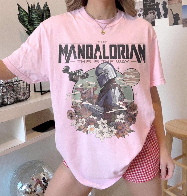May include: A light pink t-shirt featuring a graphic from The Mandalorian. The design includes the text "THE MANDALORIAN THIS IS THE WAY" above an illustration of the Mandalorian, Grogu, and floral elements. The shirt has short sleeves.