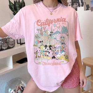 May include: Pink t-shirt featuring a Disneyland graphic with Mickey Mouse, Minnie Mouse, Donald Duck, and other characters. The shirt has the words "California" and "Disneyland" printed on it.