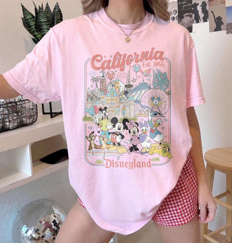 May include: Pink t-shirt featuring a Disneyland graphic with Mickey Mouse, Minnie Mouse, Donald Duck, and other characters. The shirt has the words "California" and "Disneyland" printed on it.