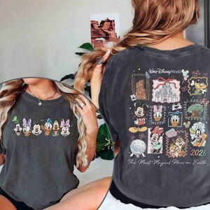 May include: Dark gray t-shirt featuring Disney characters on the front and back. The front displays a row of cartoon faces. The back has a collage of Disney World images and characters, with the text "Walt Disney World" and "2026".