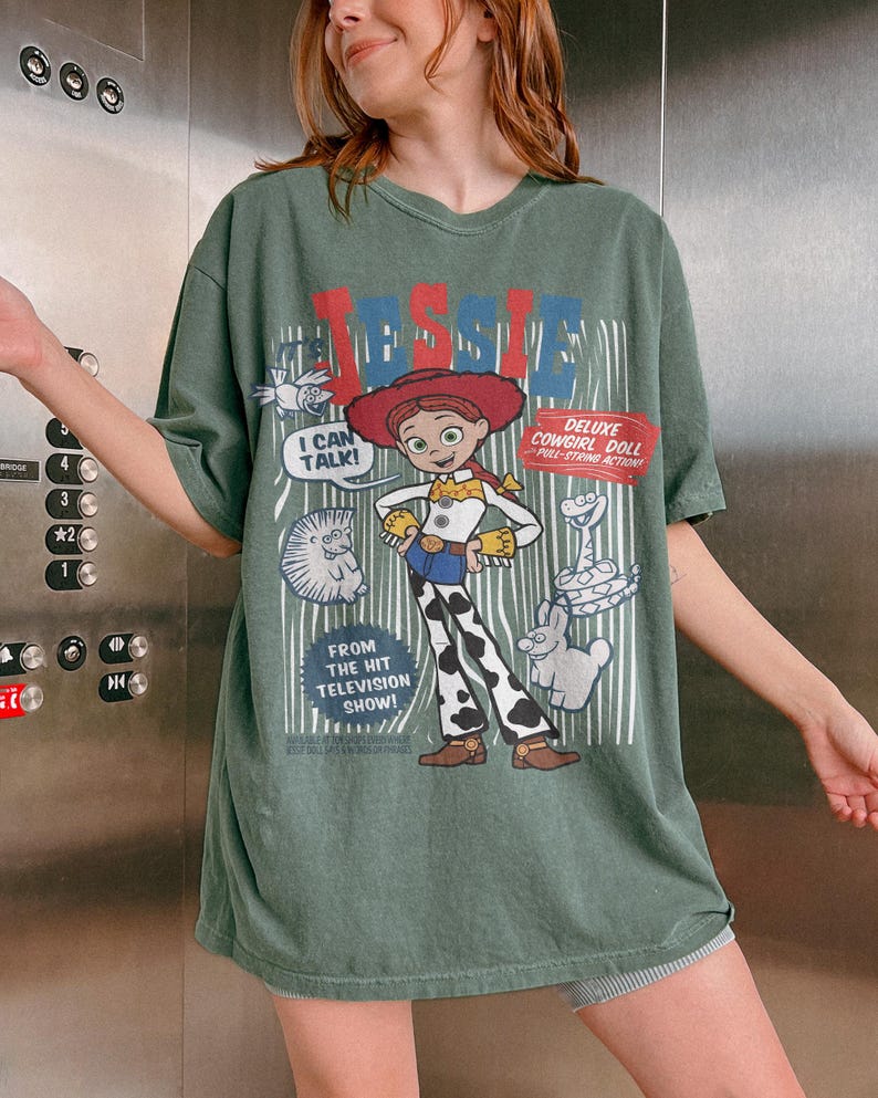 May include: Green t-shirt featuring a cartoon graphic of Jessie, the cowgirl doll, with the text "JESSIE" in red and blue. The shirt also includes the phrases "I CAN TALK!" and "DELUXE COWGIRL DOLL PULL-STRING ACTION!"