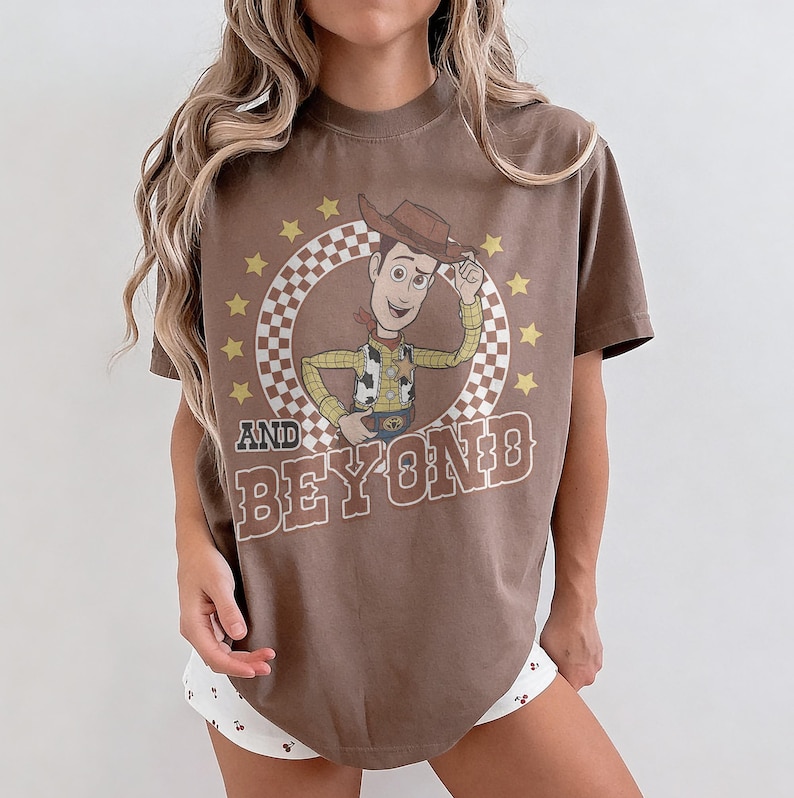 May include: A brown short-sleeve t-shirt featuring a graphic of a cowboy character in a checkered circle surrounded by yellow stars. The text "AND BEYOND" is printed in large brown letters below the character. The shirt has a crew neckline.