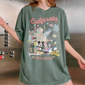 May include: Green t-shirt featuring a Disneyland graphic. The graphic includes the text "California Est. 1955" and images of Mickey Mouse, Minnie Mouse, Donald Duck, and other Disney characters. The shirt has a vintage, slightly faded look.