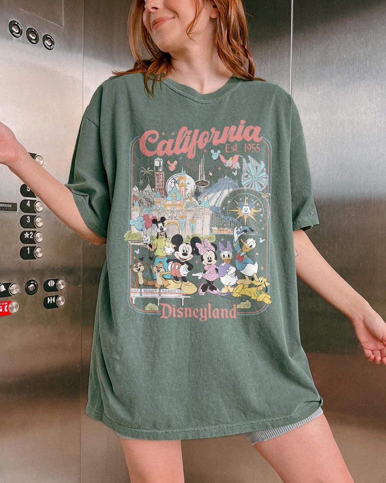 May include: Green t-shirt featuring a Disneyland graphic. The graphic includes the text "California Est. 1955" and images of Mickey Mouse, Minnie Mouse, Donald Duck, and other Disney characters. The shirt has a vintage, slightly faded look.