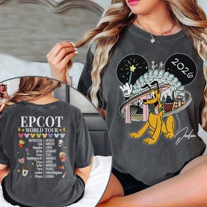 May include: Dark gray t-shirt featuring a Pluto graphic with the text "2026" and "EPCOT WORLD TOUR". The back of the shirt lists cities like Mexico City, Paris, and Tokyo. The shirt has a vintage, worn look.