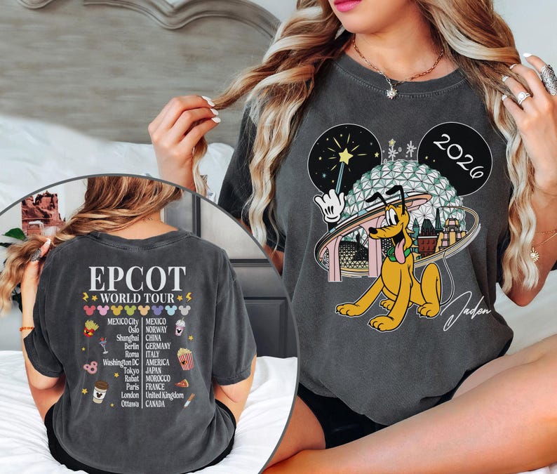 May include: Dark gray t-shirt featuring a Pluto graphic with the text "2026" and "EPCOT WORLD TOUR". The back of the shirt lists cities like Mexico City, Paris, and Tokyo. The shirt has a vintage, worn look.