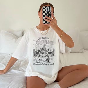 May include: White t-shirt featuring a black and white Disneyland graphic with Mickey and Minnie Mouse, a castle, and the text "CALIFORNIA Disneyland Est. 1955" and "The happiest place on earth". The shirt is oversized.