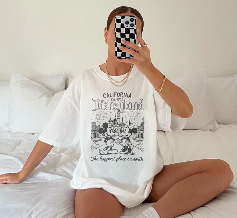 May include: White t-shirt featuring a black and white Disneyland graphic with Mickey and Minnie Mouse, a castle, and the text "CALIFORNIA Disneyland Est. 1955" and "The happiest place on earth". The shirt is oversized.