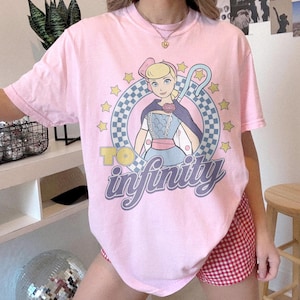 May include: A light pink short-sleeve t-shirt featuring a graphic of a blonde character with a shepherd's crook inside a blue and white checkered circle. The text "TO infinity" is printed in yellow and purple below the character, surrounded by yellow stars.