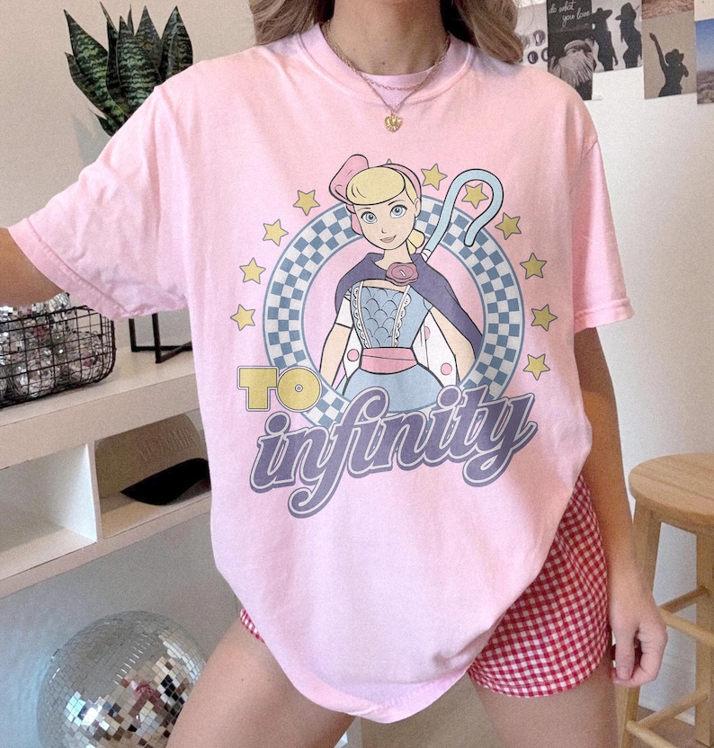 May include: A light pink short-sleeve t-shirt featuring a graphic of a blonde character with a shepherd's crook inside a blue and white checkered circle. The text "TO infinity" is printed in yellow and purple below the character, surrounded by yellow stars.