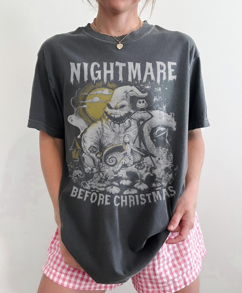 May include: Dark gray t-shirt featuring a graphic from "The Nightmare Before Christmas." The design includes the text "NIGHTMARE BEFORE CHRISTMAS" above and below a detailed illustration of characters and scenes from the movie. The shirt is worn with pink and white checkered shorts.