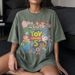 May include: A faded olive green short-sleeve t-shirt featuring a graphic print of various Toy Story characters. The center of the design displays the text Disney Pixar Toy Story 5 in yellow and red lettering, surrounded by illustrations of the movie cast.