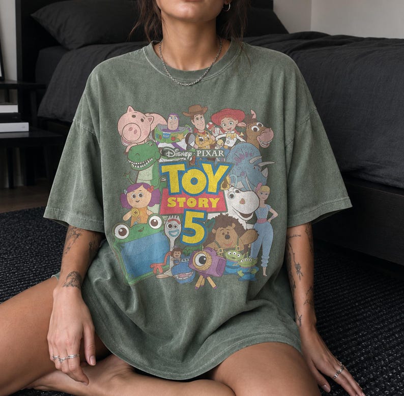May include: A faded olive green short-sleeve t-shirt featuring a graphic print of various Toy Story characters. The center of the design displays the text Disney Pixar Toy Story 5 in yellow and red lettering, surrounded by illustrations of the movie cast.