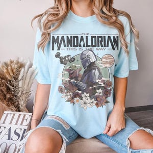 May include: Light blue t-shirt featuring a graphic of The Mandalorian and Grogu, with the text "THE MANDALORIAN - THIS IS THE WAY." The design includes a space ship, a planet, and floral elements.