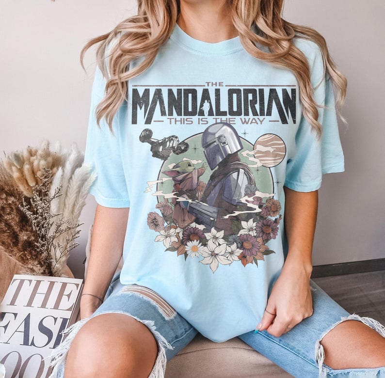 May include: Light blue t-shirt featuring a graphic of The Mandalorian and Grogu, with the text "THE MANDALORIAN - THIS IS THE WAY." The design includes a space ship, a planet, and floral elements.