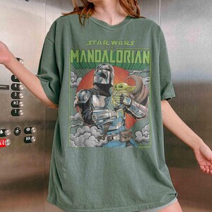 May include: Green t-shirt with a Star Wars: The Mandalorian graphic. The design shows the Mandalorian with Grogu, set against a red circle and clouds. The text "STAR WARS THE MANDALORIAN" is at the top of the image.
