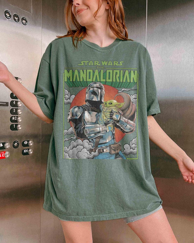 May include: Green t-shirt with a Star Wars: The Mandalorian graphic. The design shows the Mandalorian with Grogu, set against a red circle and clouds. The text "STAR WARS THE MANDALORIAN" is at the top of the image.