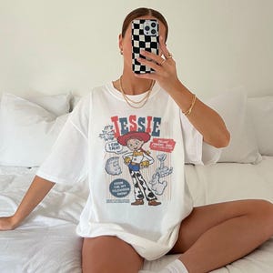 May include: White t-shirt featuring a graphic of the character Jessie from the animated film Toy Story. The design includes the name "Jessie" in red and blue, along with other cartoon elements. The shirt is oversized.