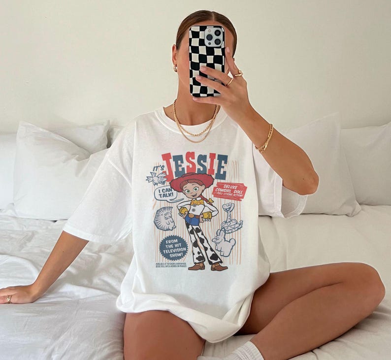 May include: White t-shirt featuring a graphic of the character Jessie from the animated film Toy Story. The design includes the name "Jessie" in red and blue, along with other cartoon elements. The shirt is oversized.