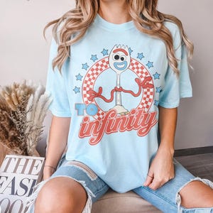 May include: A light blue short-sleeve t-shirt featuring a graphic of the character Forky inside a red and white checkered circle. The design includes blue stars and the red text "TO infinity" printed below the character on the front of the shirt.