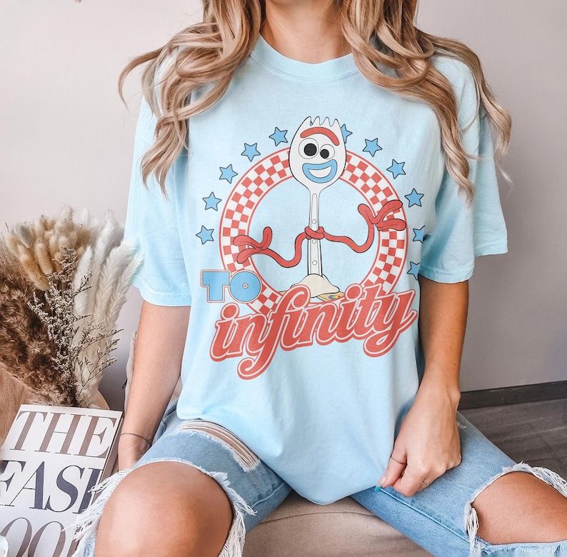 May include: A light blue short-sleeve t-shirt featuring a graphic of the character Forky inside a red and white checkered circle. The design includes blue stars and the red text "TO infinity" printed below the character on the front of the shirt.