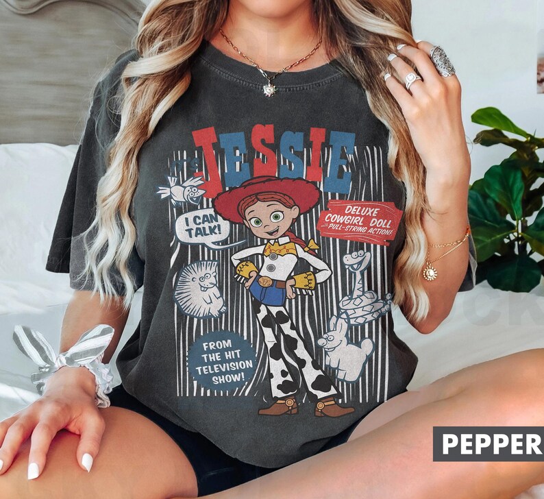 May include: Dark gray t-shirt featuring a cartoon image of Jessie, the cowgirl doll, with the text "JESSIE" above her. The shirt also includes the phrases "I CAN TALK!" and "DELUXE COWGIRL DOLL PULL-STRING ACTION."