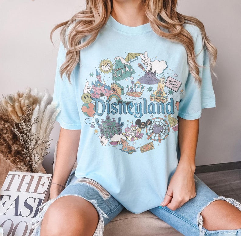 May include: Light blue t-shirt with a colorful Disneyland graphic. The design includes various park icons, the Disneyland logo, and a retro aesthetic. The shirt is a comfortable, casual style.
