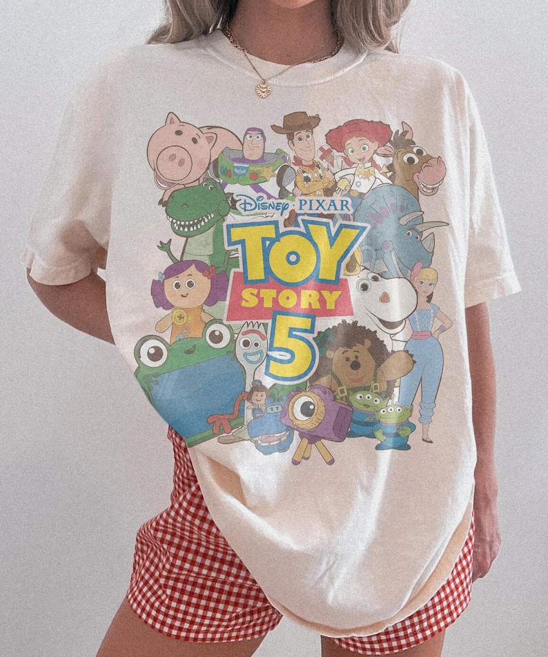 May include: An off-white short-sleeve t-shirt featuring a colorful graphic print of various Toy Story characters and the text Disney Pixar Toy Story 5. The shirt is paired with red and white gingham patterned shorts.