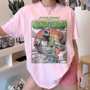 May include: Pale pink t-shirt with a graphic from "The Mandalorian." The design shows the Mandalorian character holding Grogu, with text above. The artwork is set against a red circle and green text.