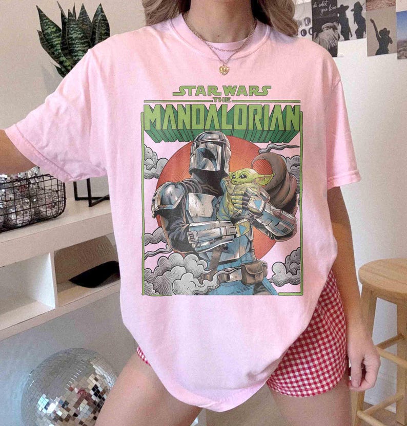 May include: Pale pink t-shirt with a graphic from "The Mandalorian." The design shows the Mandalorian character holding Grogu, with text above. The artwork is set against a red circle and green text.