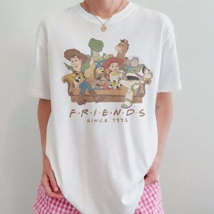 May include: A white short-sleeve t-shirt featuring a graphic print of Toy Story characters sitting on a couch. The text below the characters reads F.R.I.E.N.D.S SINCE 1995 in brown lettering. The shirt has a crew neckline and a relaxed fit.