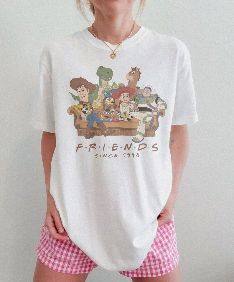May include: A white short-sleeve t-shirt featuring a graphic print of Toy Story characters sitting on a couch. The text below the characters reads F.R.I.E.N.D.S SINCE 1995 in brown lettering. The shirt has a crew neckline and a relaxed fit.