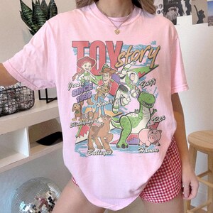 May include: A light pink short-sleeve t-shirt featuring a graphic print of Toy Story characters including Jessie, Woody, Buzz Lightyear, Slinky Dog, Bullseye, Rex, and Hamm. The text reads Toy Story, Jessie Since 1995, Woody, Buzz, Slinky Dog, Bullseye, Rex, and Hamm.