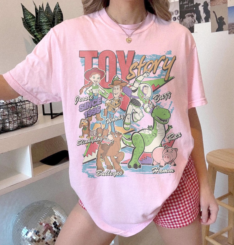May include: A light pink short-sleeve t-shirt featuring a graphic print of Toy Story characters including Jessie, Woody, Buzz Lightyear, Slinky Dog, Bullseye, Rex, and Hamm. The text reads Toy Story, Jessie Since 1995, Woody, Buzz, Slinky Dog, Bullseye, Rex, and Hamm.