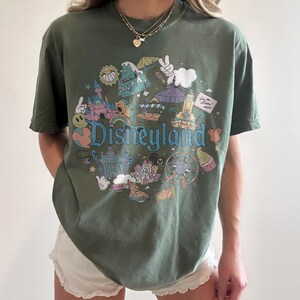 May include: Olive green t-shirt with a colorful Disneyland graphic. The design features iconic park elements like the castle, teacups, and a Ferris wheel, with the word "Disneyland" in a playful font.