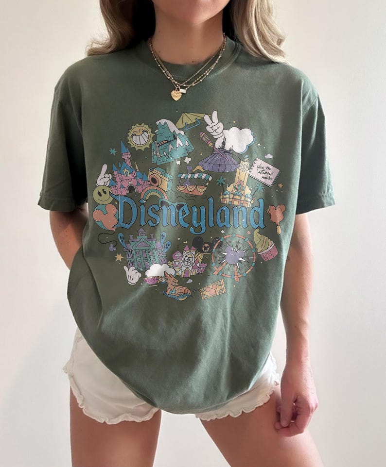 May include: Olive green t-shirt with a colorful Disneyland graphic. The design features iconic park elements like the castle, teacups, and a Ferris wheel, with the word "Disneyland" in a playful font.